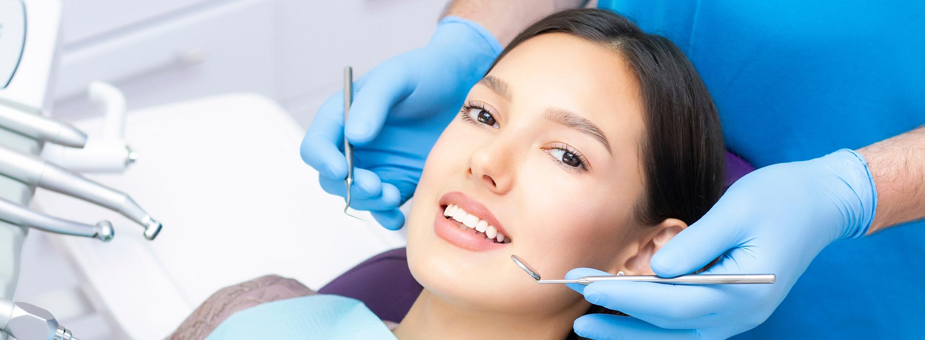 The image shows a person receiving dental care, with a dentist performing a procedure using a drill while the patient is seated in a dental chair, wearing protective eyewear, and smiling.