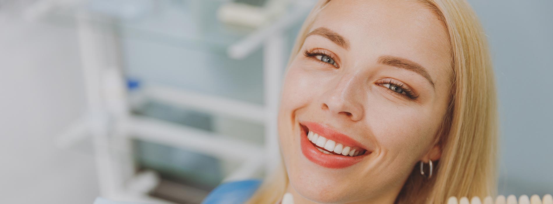 The image features a smiling woman with red lipstick, looking directly at the camera, set against a blurred background that appears to be an indoor setting with shelves containing various items.