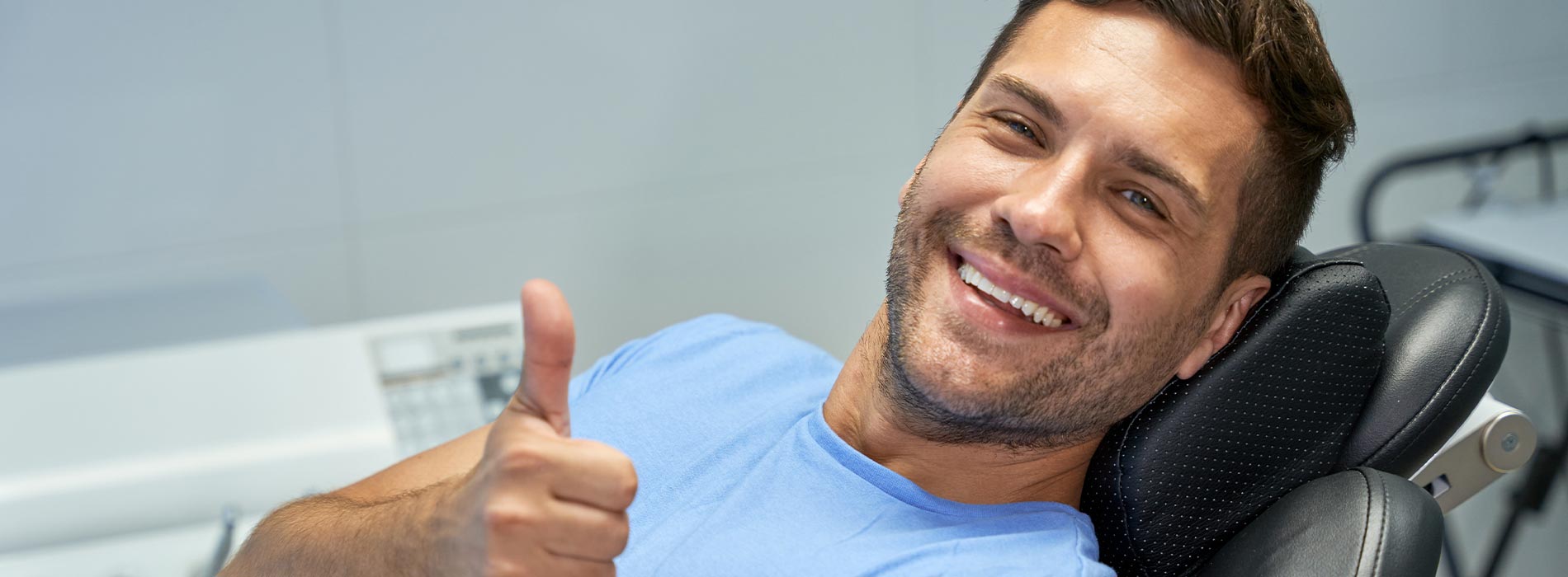 A man sitting in a dental chair, giving a thumbs up with his left hand.