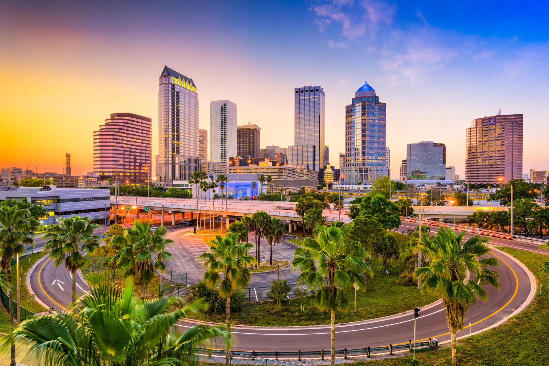 The image depicts a city skyline at sunset with palm trees and a curved road leading towards a large, illuminated cityscape.