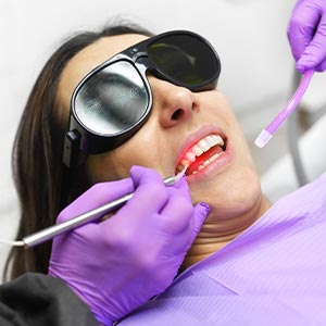 The image shows two photos of a woman receiving dental treatment  she is wearing protective eyewear and has her mouth open while sitting in a dentist s chair, with visible dental tools and equipment around her.