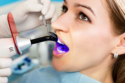 The image depicts a woman sitting in a dental chair with her mouth open, receiving a dental cleaning or examination from a professional using a dental mirror and an instrument.