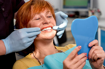 A woman sitting in a dental chair with a blue mouthguard, smiling at the camera while holding a toothbrush, with a dental hygienist adjusting her teeth using a mirror and tweezers.
