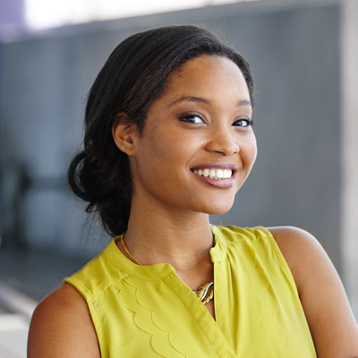 The image shows a woman with long hair smiling at the camera, wearing a yellow top and standing against a backdrop that includes a metal structure.