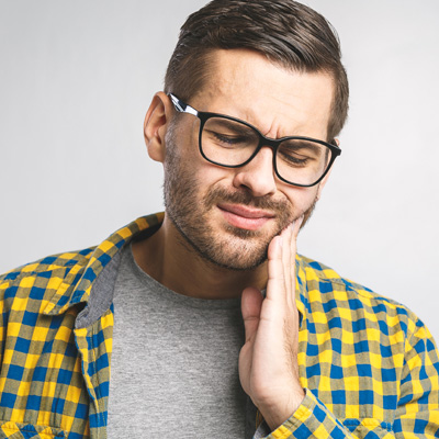 The image shows a man with glasses and a beard wearing a yellow plaid shirt, appearing to be in discomfort or pain, possibly due to dental issues, with his hand on his chin.