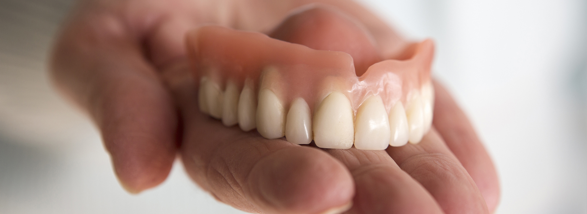 The image shows a person holding an open mouth with artificial teeth, which appear to be dentures, against a plain background.