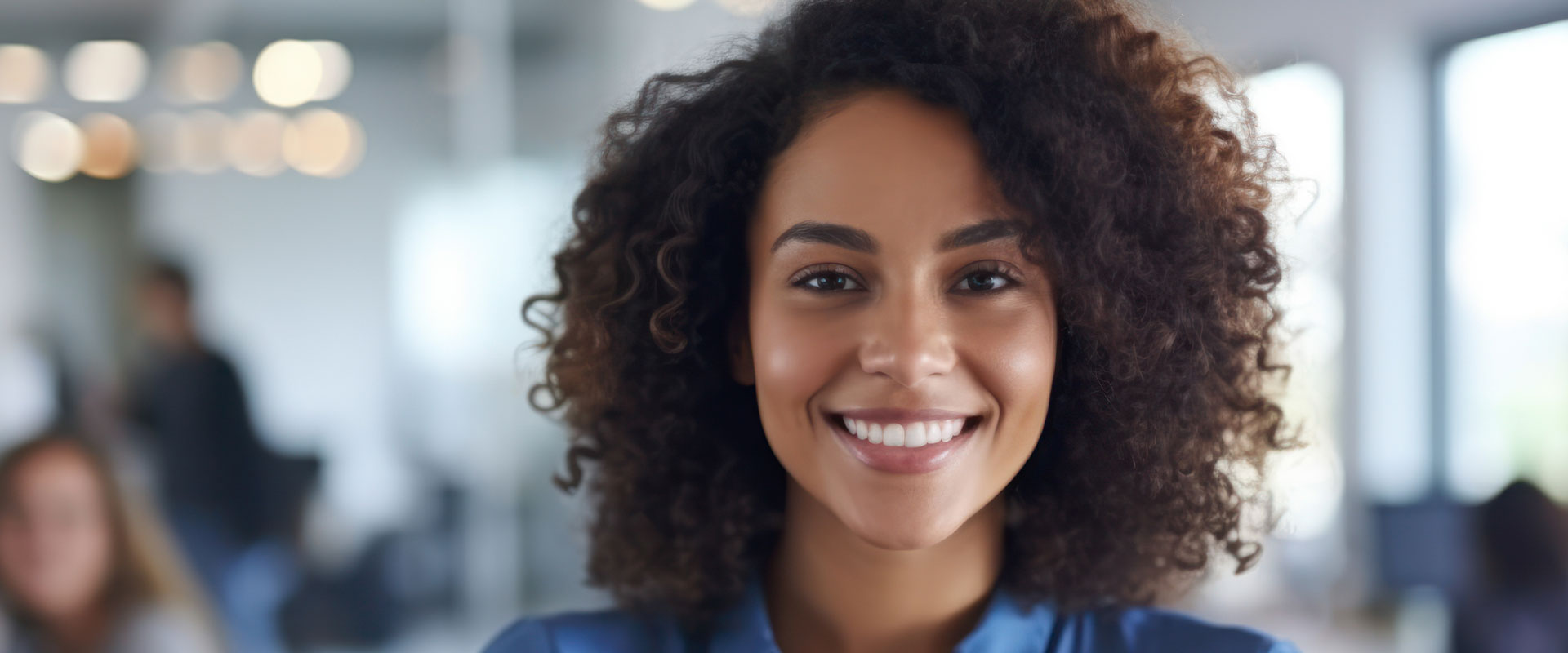 This is a photograph of a person with curly hair smiling at the camera, wearing a dark top and seated indoors, possibly in an office setting.