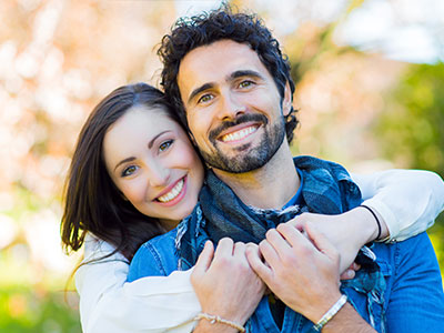 The image shows a man and woman embracing each other with warm smiles, both wearing casual clothing, set against an outdoor backdrop with trees and foliage.