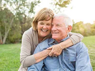 An elderly couple embracing each other outdoors, with a man wearing a blue shirt and a woman in a white top.