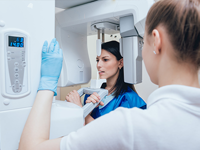 In the image, there are two individuals standing in front of a large, white, medical-looking machine with screens displaying various information. A woman is operating the machine, while a man stands nearby observing her work.