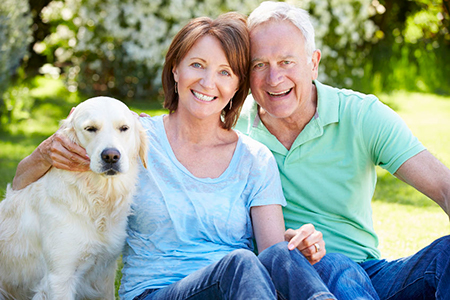 The image shows an elderly couple sitting outdoors with their golden retriever dog, posing for a photo together.