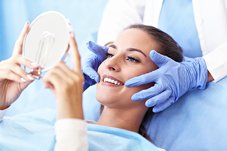 In the image, a woman is seated on a dental chair with her eyes closed, receiving a dental treatment. A dentist is holding up a mirror to show her teeth, while another person appears to be taking a photo of the moment.
