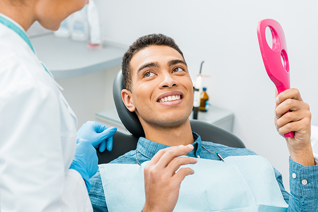 A man sitting in a dental chair with a smile while holding a pink object, receiving dental care from a professional in a white coat and mask.