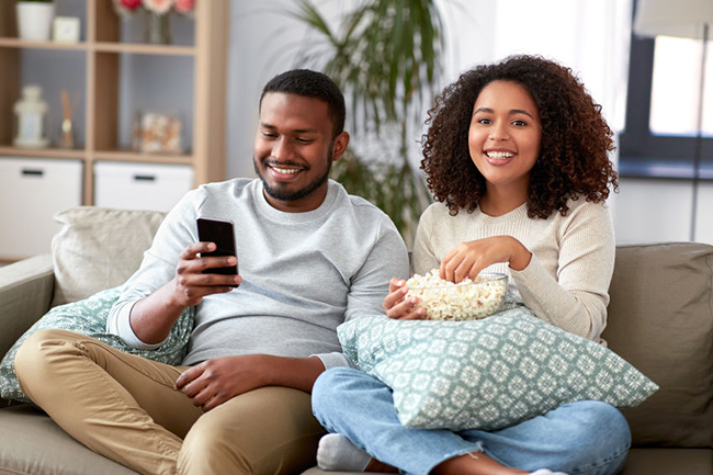 A smiling couple sitting on a couch, enjoying popcorn while watching something on their phones.