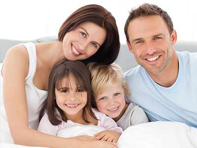 This image depicts a family of four people, including two adults and two children, smiling together on a couch with a cozy and warm atmosphere.