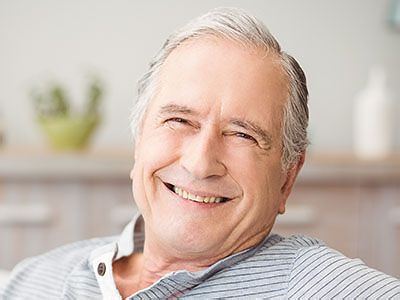 The image shows a smiling older man sitting comfortably at home, possibly enjoying a moment of relaxation or contentment.