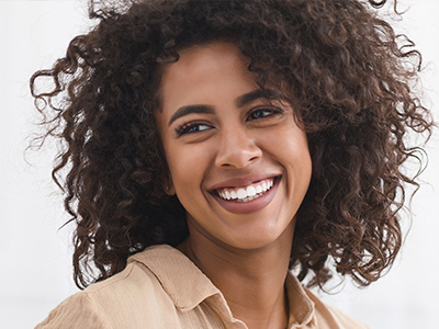 The image shows a woman with curly hair smiling at the camera  she has a radiant complexion and appears to be wearing a light-colored top, possibly a blouse or shirt.